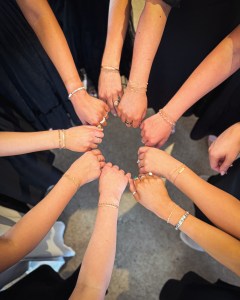 Eight hands displaying matching bracelets, arranged in a circle, against a backdrop of dark clothing.