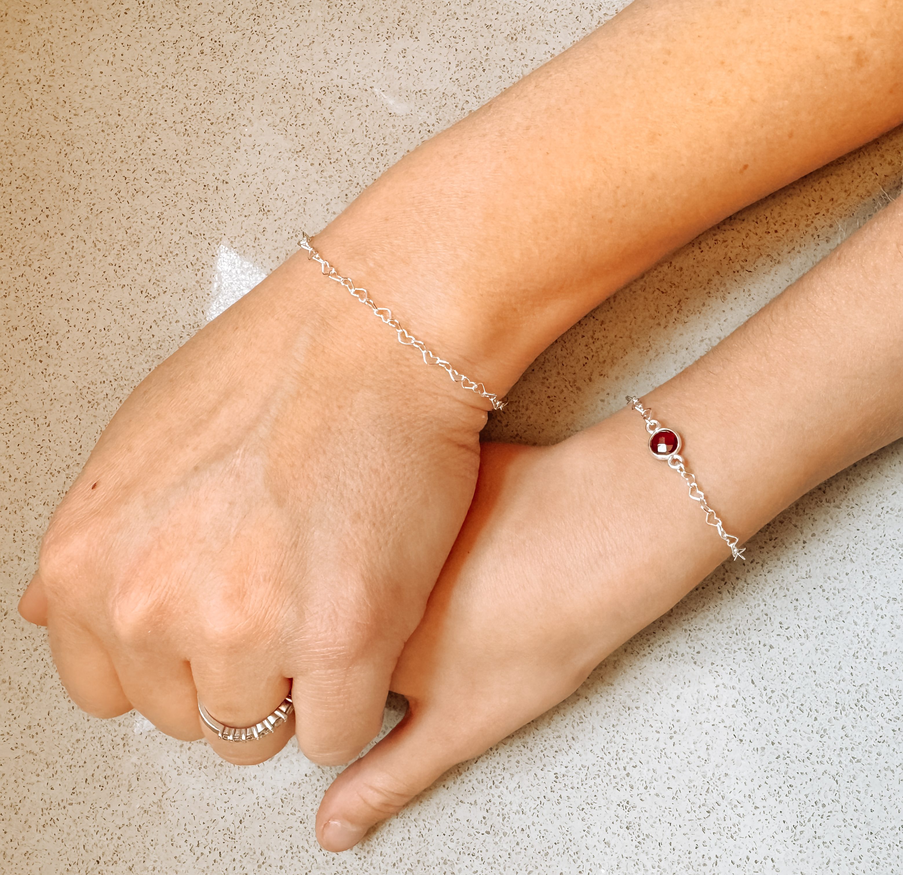 Two hands wearing silver bracelets, one featuring a red gemstone, resting on a light-colored surface.